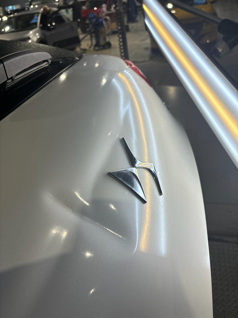 Hayon blanc enfoncé sous le logo, déformation visible dans les reflets de la lampe